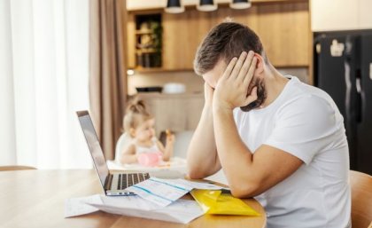 a man sits at a table with his face in his hands, in front of him are papers and a laptop, behind him is a small child in a high chair 